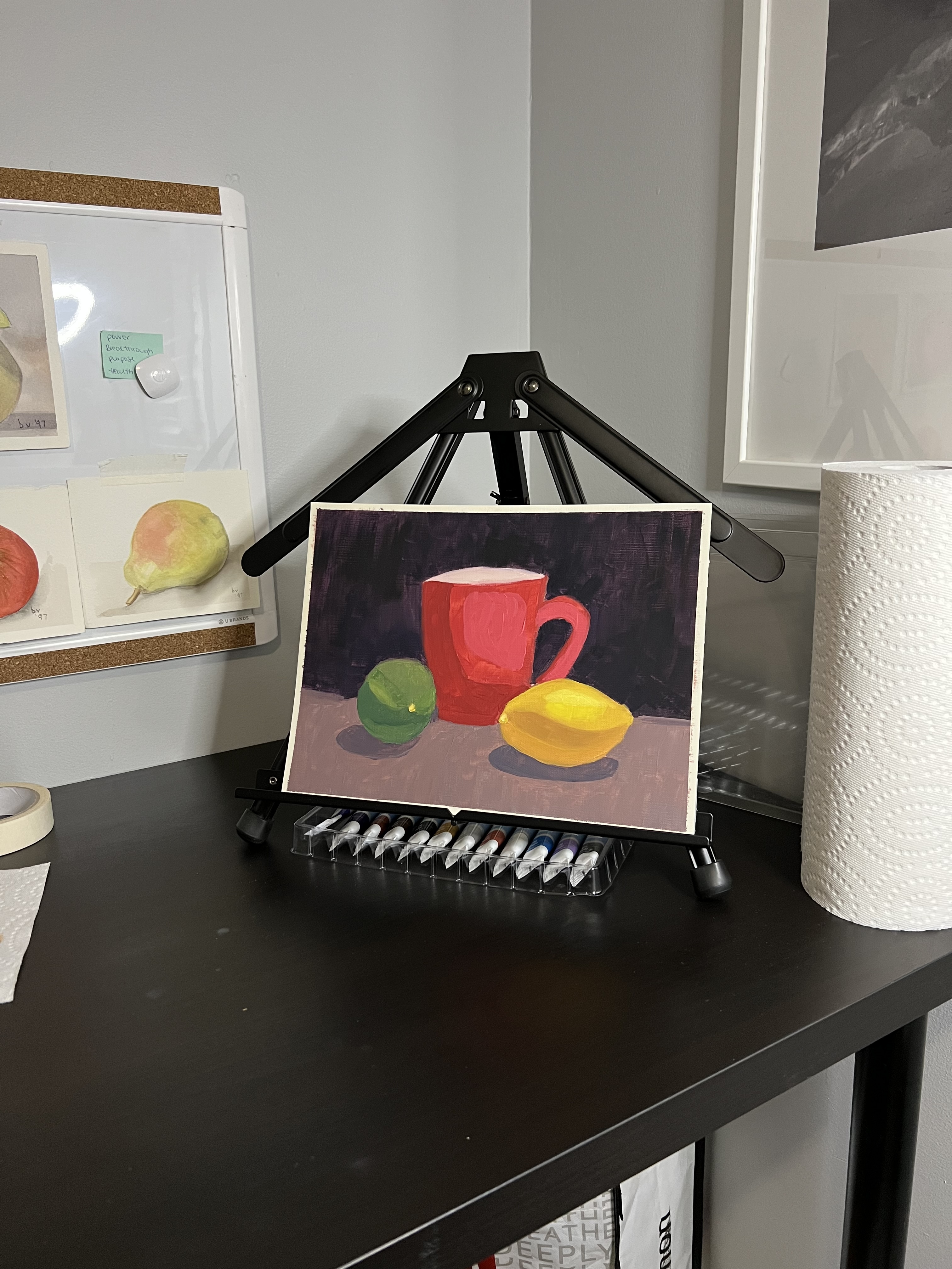 Oil painting of a red cup, lime and lemon on a table. Painting is sitting on an easel which is sitting on a table.