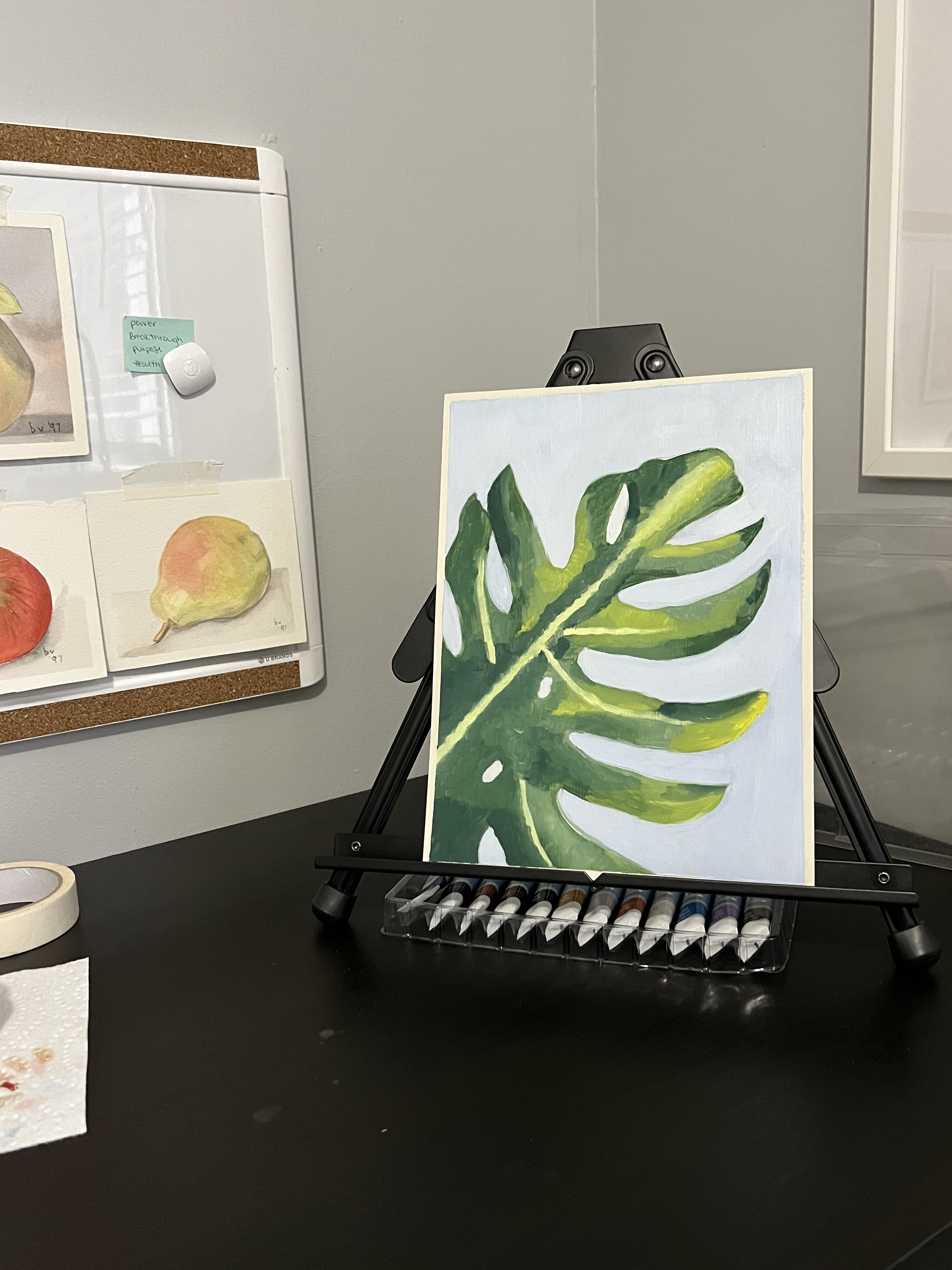 Oil painting sitting on an easel that is placed on a table. Painting is of a green monstera leaf with a light blue background