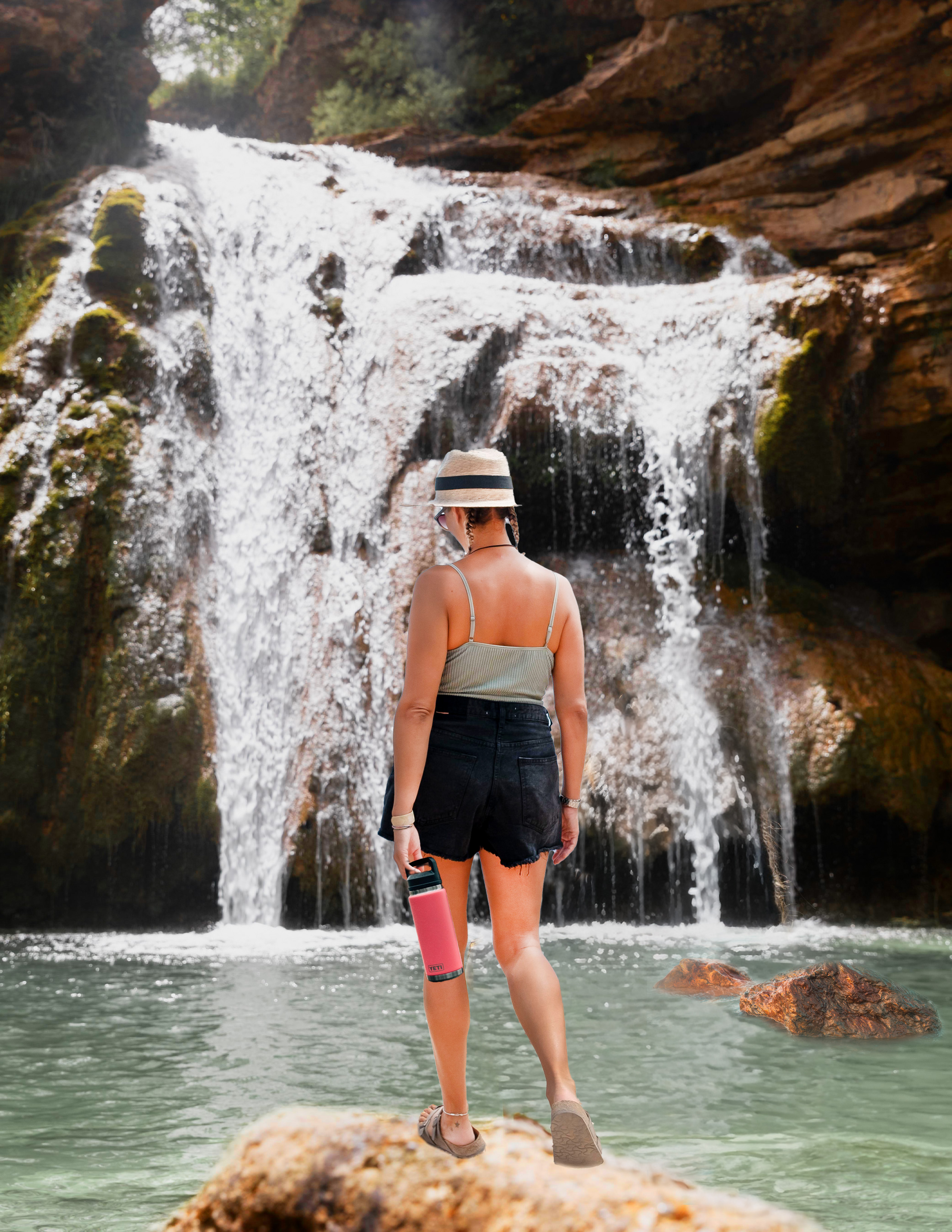 girl standing on a rock in front of a water fall. girl is holding a bright pink water bottle and looking down at the water 