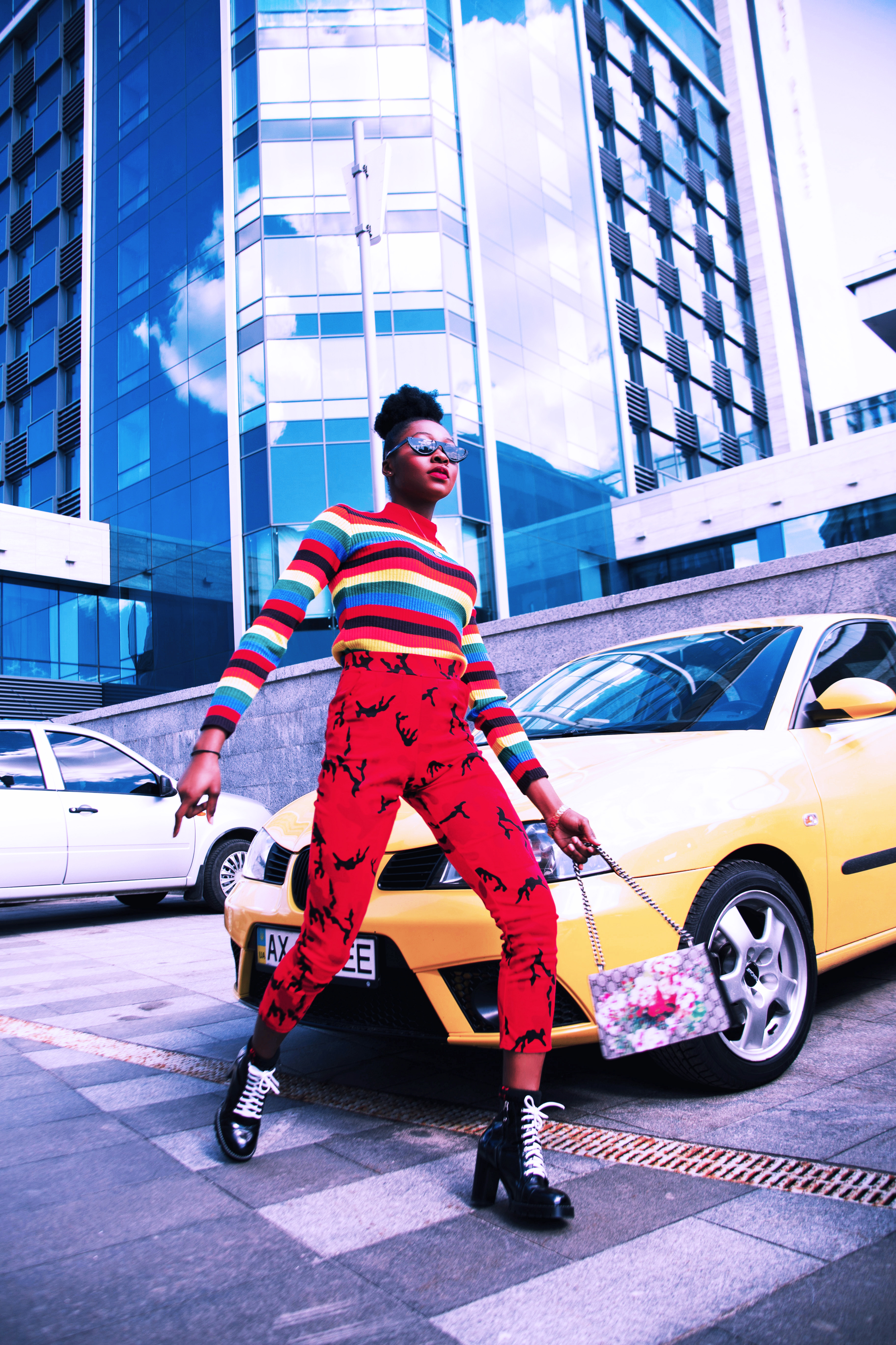 Stylish person in a colorful striped sweater and red patterned pants posing in front of a yellow car and modern city buildings.